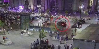 A car drives through a busy plaza in front of a cathedral, heading toward a crowd of people.