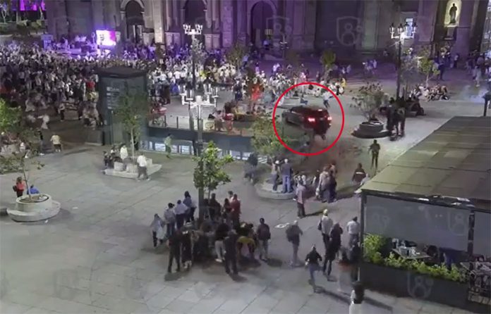 A car drives through a busy plaza in front of a cathedral, heading toward a crowd of people.