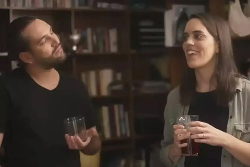 A man and a woman in their 30s holding cocktail glasses and looking at each other as they talk in a room with bookcases lining the walls.