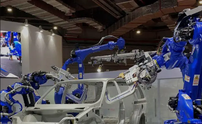 Shandong Golden Empire manufacturing demonstration with a white car frame in a conference hall and blue robotic arms on either side of the frame manipulating auto parts