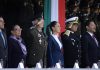 Sheinbaum stands in front of a tri-colored banner, as military officials salute at her side.