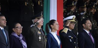 Sheinbaum stands in front of a tri-colored banner, as military officials salute at her side.