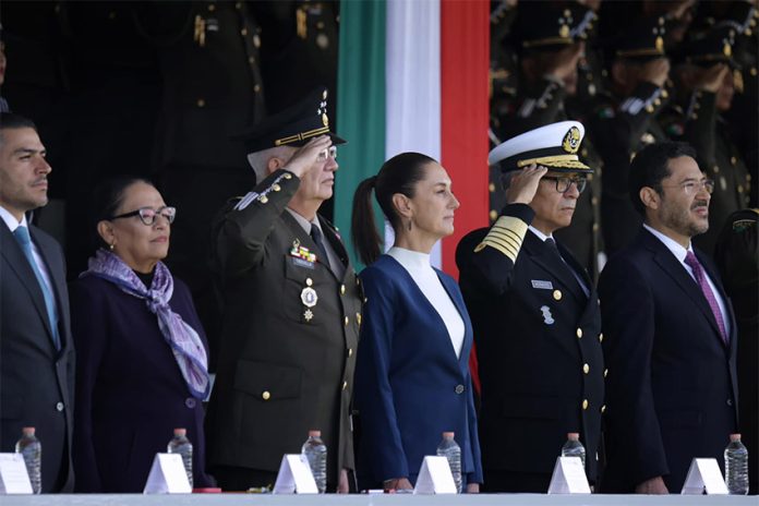 Sheinbaum stands in front of a tri-colored banner, as military officials salute at her side.