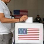 Man in a T-shirt and slacks putting a paper ballot into a box with a printout of the US flag in a room where people in the background are on laptops at a table and a U.S. flag is pinned to the wall.