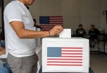 Man in a T-shirt and slacks putting a paper ballot into a box with a printout of the US flag in a room where people in the background are on laptops at a table and a U.S. flag is pinned to the wall.