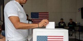 Man in a T-shirt and slacks putting a paper ballot into a box with a printout of the US flag in a room where people in the background are on laptops at a table and a U.S. flag is pinned to the wall.