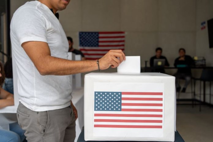 Man in a T-shirt and slacks putting a paper ballot into a box with a printout of the US flag in a room where people in the background are on laptops at a table and a U.S. flag is pinned to the wall.