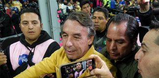 A gray-haired man pushes through a crowd of people taking pictures of him and holding microphones