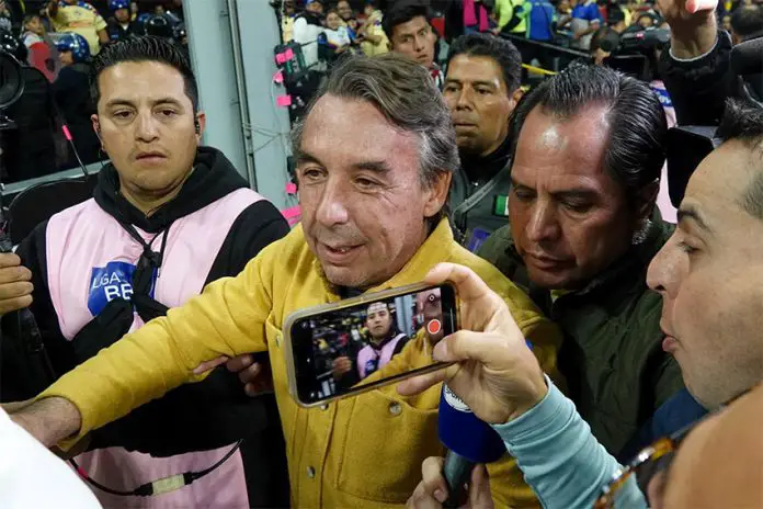 A gray-haired man pushes through a crowd of people taking pictures of him and holding microphones