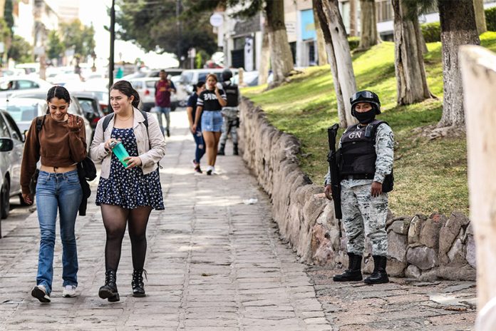 Young women walk down a sidewalk while a member of the National Guard stands guard, illustrating the level of perceived insecurity in Mexican cities