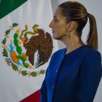 Mexico's Claudia Sheinbaum standing sideways in a blue suit jacket looking at something off-camera. Behind her is a large Mexican flag