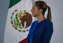 Mexico's Claudia Sheinbaum standing sideways in a blue suit jacket looking at something off-camera. Behind her is a large Mexican flag