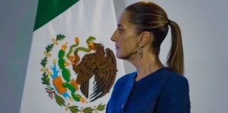 Mexico's Claudia Sheinbaum standing sideways in a blue suit jacket looking at something off-camera. Behind her is a large Mexican flag