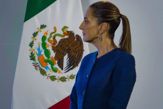 Mexico's Claudia Sheinbaum standing sideways in a blue suit jacket looking at something off-camera. Behind her is a large Mexican flag