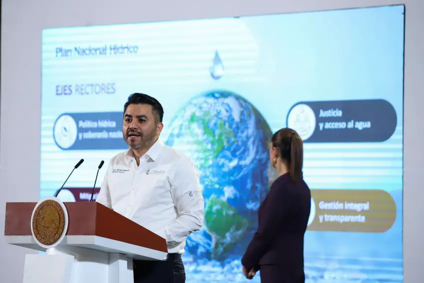 Mexico's National Water Commission (Conagua) General Director Efraín Morales López standing at a podium at a presidential press conference speaking to reporters in front of a projection of a report on the National Water Plan. President Sheinbaum is looking at the screen turned away from the camera