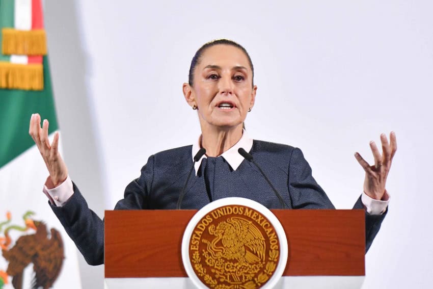 Mexico's President Claudia Sheinbaum in front of the presidential podium in the National Palace at a press conference. She is gesturing with both hands out as she speaks to reporters.