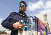 Accordion player with the popular Mexican band Buyuchek posing in sunglasses with his instrument outside in a residential area, with a blue sky and a few clouds above him.