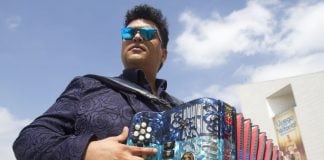 Accordion player with the popular Mexican band Buyuchek posing in sunglasses with his instrument outside in a residential area, with a blue sky and a few clouds above him.