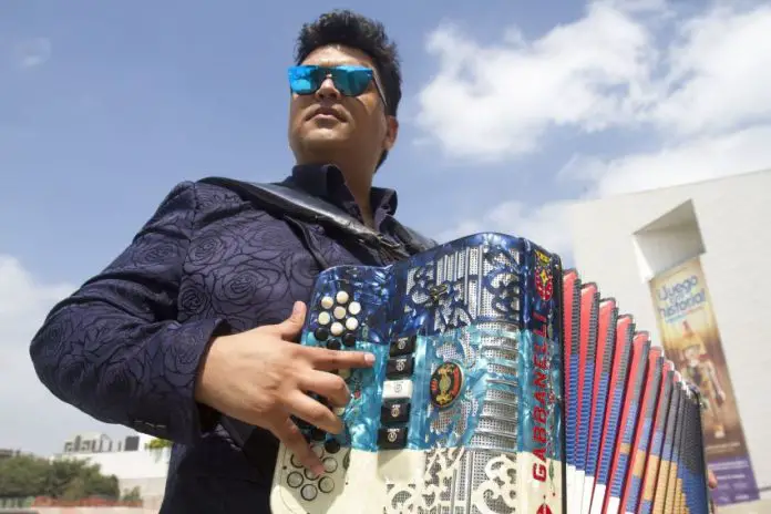 Accordion player with the popular Mexican band Buyuchek posing in sunglasses with his instrument outside in a residential area, with a blue sky and a few clouds above him.