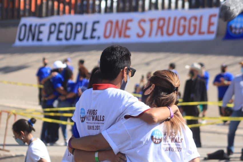 A man and woman stand backs to the camera, wearing Hugs Not Walls event T-shirts and looking at a channelized section of the Rio Grande river along the Mexico-US border.