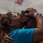 A woman in a blue shirt and temporary wrist band kisses a younger man with sun glasses on the forehead, with an American flag waving in the background.