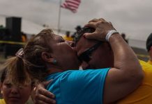 A woman in a blue shirt and temporary wrist band kisses a younger man with sun glasses on the forehead, with an American flag waving in the background.