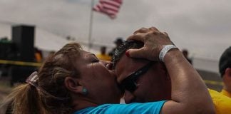 A woman in a blue shirt and temporary wrist band kisses a younger man with sun glasses on the forehead, with an American flag waving in the background.