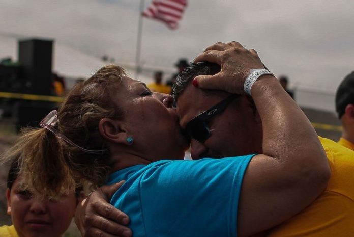 A woman in a blue shirt and temporary wrist band kisses a younger man with sun glasses on the forehead, with an American flag waving in the background.