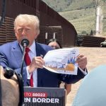 Donald Trump holding a printout of a color line graph whose details are not visible while he stands at a podium in a desert location near the U.S. border.