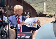 Donald Trump holding a printout of a color line graph whose details are not visible while he stands at a podium in a desert location near the U.S. border.