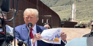 Donald Trump holding a printout of a color line graph whose details are not visible while he stands at a podium in a desert location near the U.S. border.