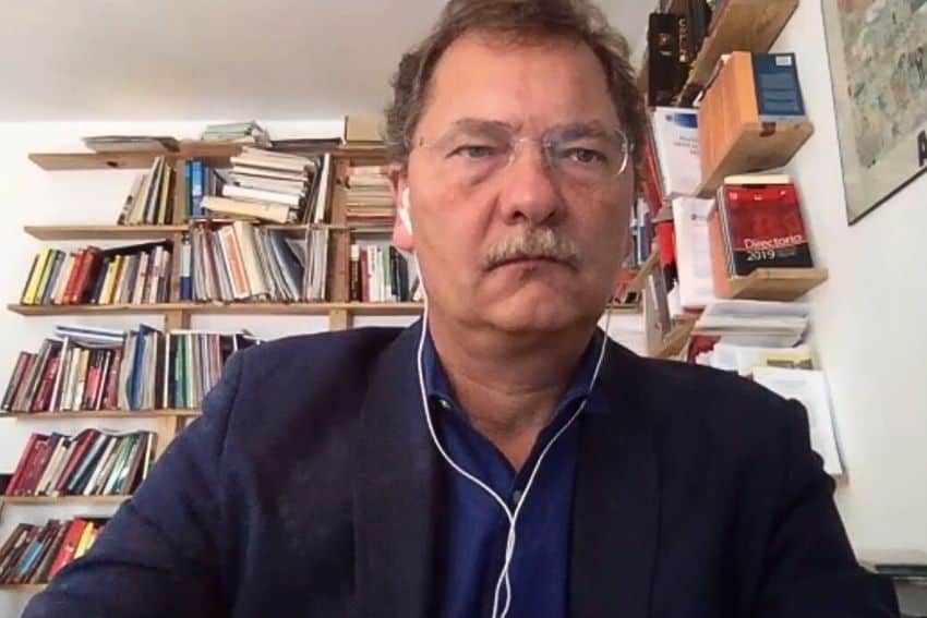 Enrique Dussel Peters on a videoconference call in his home. In the background is full wall bookshelf crammed with books.