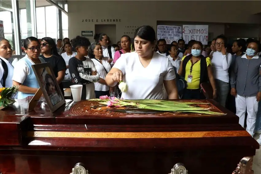 A woman places a white rose on top of a closed wooden coffin atop which are long-stemmed pink flowers and a framed photo of a young Mexican woman.