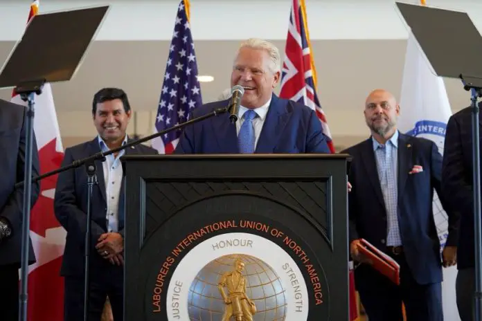 Ontario, Canada Prime Minister Doug Ford standing at a podium with the logo of the Labourers International Union of North America giving a speech. Behind him are two men watching on