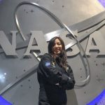 A young woman wearing a NASA jumper poses in front of a massive NASA logo