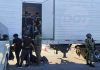 Tractor trailer with undocumented male migrants inside being ordered out by a man with a National Immigration Institute uniform. Near him are other migrants already outside the truck and a Mexican soldier in a military gear and holding an automatic weapon stands guard.