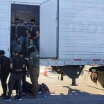 Tractor trailer with undocumented male migrants inside being ordered out by a man with a National Immigration Institute uniform. Near him are other migrants already outside the truck and a Mexican soldier in a military gear and holding an automatic weapon stands guard.