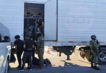 Tractor trailer with undocumented male migrants inside being ordered out by a man with a National Immigration Institute uniform. Near him are other migrants already outside the truck and a Mexican soldier in a military gear and holding an automatic weapon stands guard.