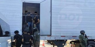 Tractor trailer with undocumented male migrants inside being ordered out by a man with a National Immigration Institute uniform. Near him are other migrants already outside the truck and a Mexican soldier in a military gear and holding an automatic weapon stands guard.