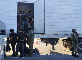 Tractor trailer with undocumented male migrants inside being ordered out by a man with a National Immigration Institute uniform. Near him are other migrants already outside the truck and a Mexican soldier in a military gear and holding an automatic weapon stands guard.