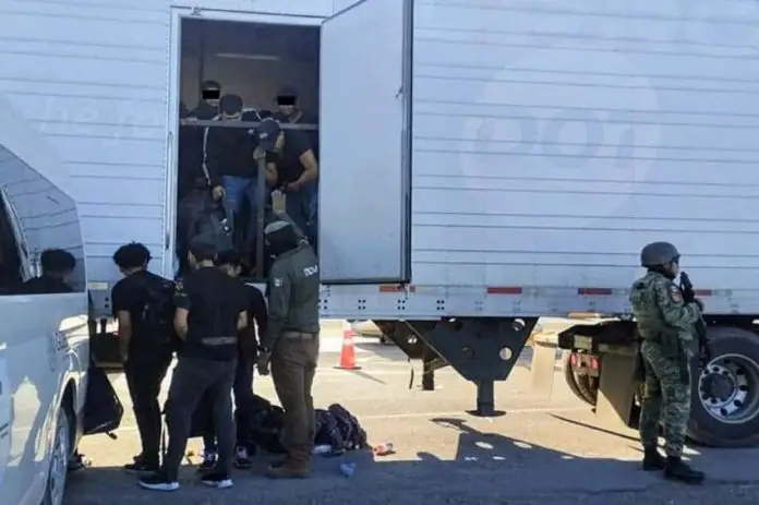 Tractor trailer with undocumented male migrants inside being ordered out by a man with a National Immigration Institute uniform. Near him are other migrants already outside the truck and a Mexican soldier in a military gear and holding an automatic weapon stands guard.