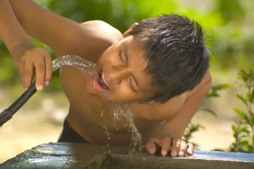 A boy happily drinking water directly from a coursing water hose