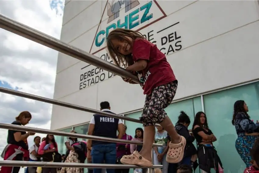 Migrant girl around 10 years old playing on a railing as nearby migrants stand around outside the Zacatecas, Mexico state human rights commission.