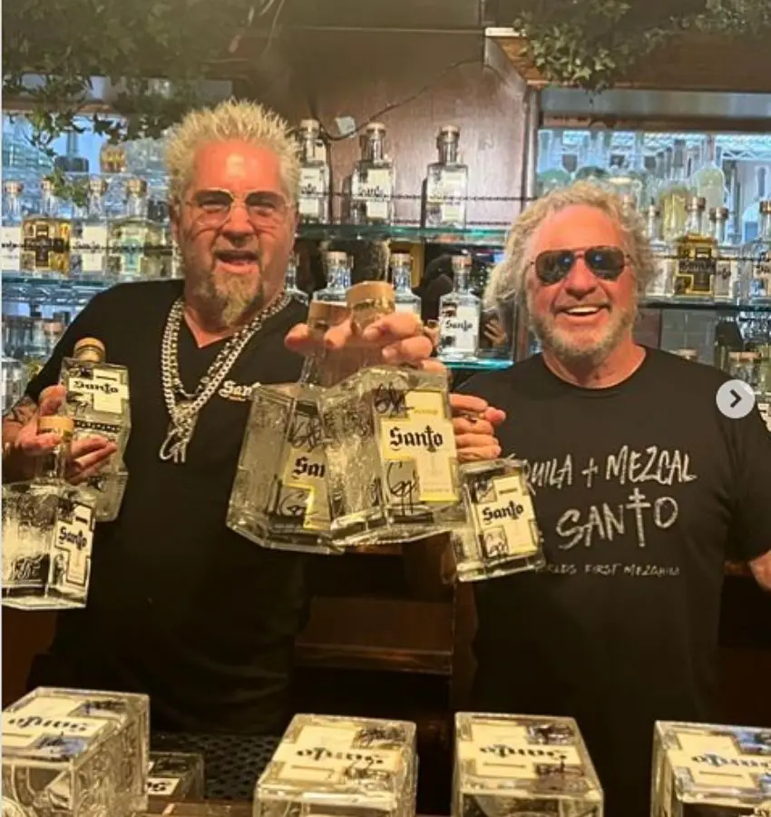 Guy Fieri and Sammy Hagar posing with bottles of their Santo brand liquor and smiling, surrounded by bar shelves holding bottles of the brand.