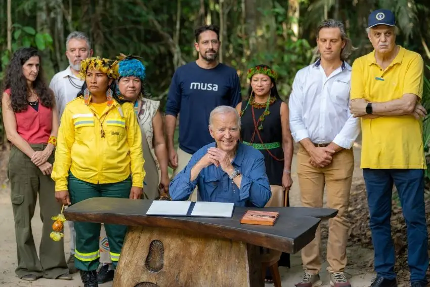 Presiden AS Joe Biden duduk di meja kayu pedesaan di hutan hujan Amazon dikelilingi oleh sekelompok kecil aktivis lingkungan. Di atas meja di depannya ada portofolio dengan dokumen yang harus ditandatangani dan pena tertutup yang tampak mahal di dalam kotak kayu.