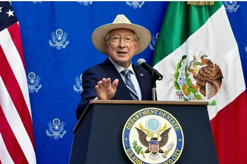 US Ambassador Ken Salazar standing at a podium with the US and Mexico flags behind him as well as a blue curtained wall that has a repeated pattern of the logo for the US Department of State.