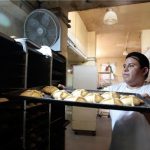 Mexican man in a white baker's tee shirt and apron puts a baking sheet of bread rolls onto an industrial-sized rack