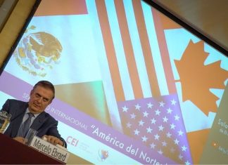 Mexico's Economy Minister Marcelo Ebrard sitting at a panel discussion table onstage with his name placard in front of him and a jug of water. Behind him is a projection screen with images of the Mexican, Canadian and US flags and Spanish words saying "International Seminar: North America: What lies ahead"