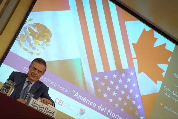 Mexico's Economy Minister Marcelo Ebrard sitting at a panel discussion table onstage with his name placard in front of him and a jug of water. Behind him is a projection screen with images of the Mexican, Canadian and US flags and Spanish words saying 