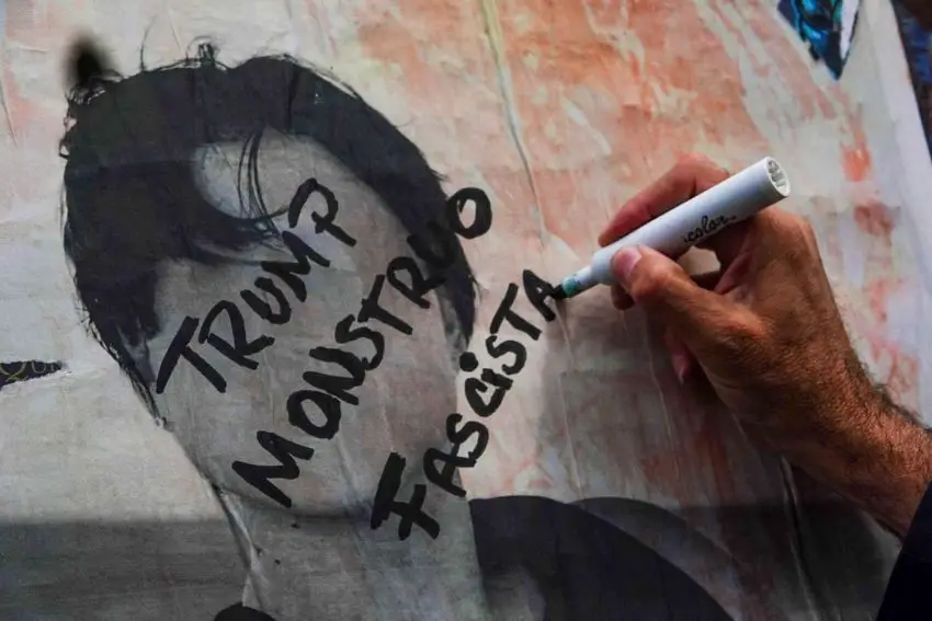 Person's hand writing with a dry-erase marker on a painting of a faceless man. Written on the figure's face in Spanish are the words "Trump" "Monster" and "Fascist."